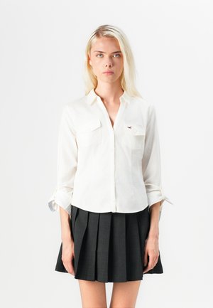 White button-up shirt with two front pockets and rolled sleeves, paired with a black pleated skirt. Cotton fabric with a smooth texture.