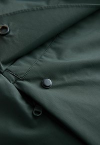 Dark green fabric with a smooth texture features a button and zipper detail, showcasing a folded section and subtle ribbed pattern.