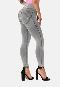 Nainen, joka pukeutuu harmaisiin kapeisiin farkkuihin ja mustiin korkeakorkoisiin sandaaleihin, seisoo sivuttain valkoista taustaa vasten.