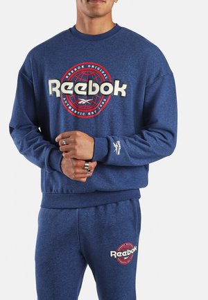 Uomo che indossa una felpa blu Reebok e pantaloni della tuta abbinati con logo rosso e bianco, mani intrecciate in vita.
