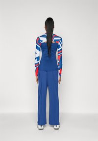 Modré dlhé nohavice so voľným strihom, v páre s priliehavým topom s farebným abstraktným vzorom v červenej a modrej farbe. Hladká textúra, minimálne detaily.