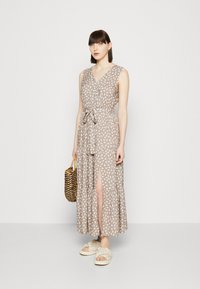 Beige ärmelloses Maxikleid mit weißen Blattmustern, V-Ausschnitt, tailliertem Gürtel und seitlichem Schlitz. Kombiniert mit hellen Plateauschuhen und einer geflochtenen Tasche.