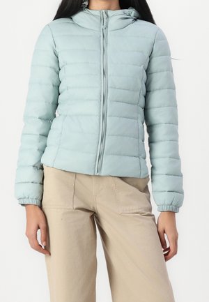 Kobieta ubrana w jasno niebieską pikowaną kurtkę puchową z kapturem, w połączeniu z beżowymi, wysokim stanem spodniami z przednimi kieszeniami.