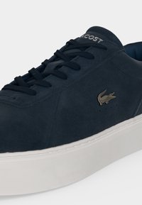 Σκούρο μπλε αθλητικό παπούτσι Lacoste με μαύρα κορδόνια, λευκή σόλα και μεταλλικό λογότυπο κροκόδειλου στο πλάι.