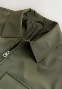 Olivengrøn jakke med spids krave, sort frontlynlås og synlige syninger ved brystlommen, lagt fladt på en lys baggrund.