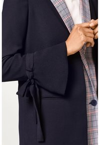 Lähellä kuva henkilöstä, joka säätää tummansinistä takkia, jossa on solmittu hiha ja ruutukuvioinen sisävuori valkoisen paidan päällä.