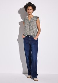 Kukkakuvioinen, vaaleanvihreä paita, jossa on frilloja hihoissa, yhdistetty tummansinihin korkeavyötäröisiin farkkuihin. Malli seisoo kädet taskuissa.