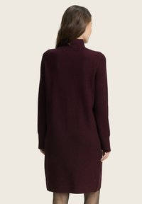 Bordeauxfarbiges Strickkleid mit hohem Kragen und langen Ärmeln, ausgestattet mit einem Schlüssellochdetail am Rücken und seitlichen Schlitzen für Bewegungsfreiheit.