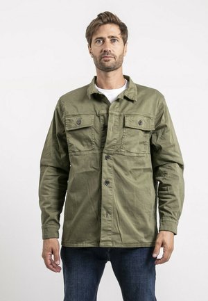 Homme aux cheveux châtain clair et barbe portant une veste boutonnée vert olive par-dessus une chemise blanche et un jean bleu foncé, debout devant un fond blanc.