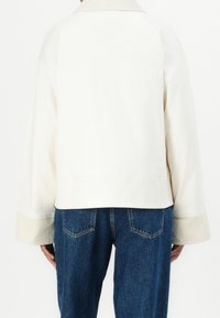 Personne debout portant une veste blanche à manches longues avec des poignets de couleur claire et un jean bleu, vue de dos.