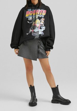 Sudadera negra con capucha que presenta un gráfico colorido de "Boruto", combinada con una minifalda gris de rayas con detalles de cordones y botines negros hasta el tobillo.