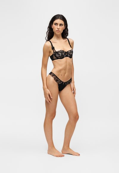 Jeune femme aux longs cheveux foncés portant un soutien-gorge en dentelle noire et un slip assorti, debout pieds nus contre un fond clair uni.