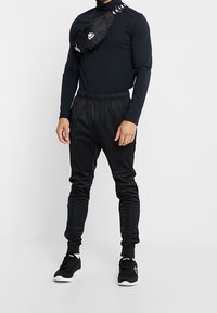 Czarna koszula z długim rękawem w zestawie z dopasowanymi czarnymi joggerami, obie z gładkiego materiału. Całość uzupełnia czarna torba na ramię z detalem logo.