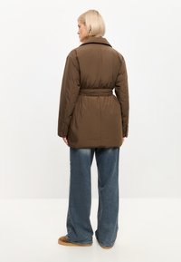 Chaqueta acolchada marrón con cinturón en la cintura, presenta un cuello clásico y puños abotonados, combinada con jeans de pierna ancha azules y zapatos marrones.