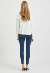 Kvinna med långt blont hår som bär en vit tröja, blå skinnyjeans och svarta klacksandaler, stående vänd bortåt mot en vit bakgrund.