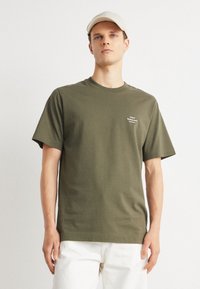 Olivovo zelený bavlnený tričko s krátkymi rukávmi a okrúhlym výstrihom, s malým bielym logom na ľavej hrudi. Model má na sebe svetlé nohavice.