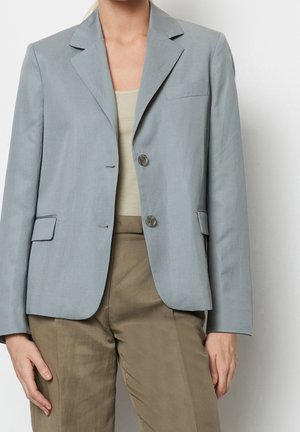 Kobieta w jasnoniebieskiej dopasowanej marynarce na dwa guziki, na beżowej prążkowanej bluzce i oliwkowozielonych spodniach z wysokim stanem, stojąca na białym tle.