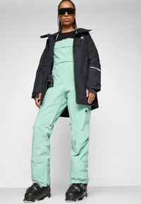 Conjunto de esquí que incluye una chaqueta negra y un mono verde menta. La chaqueta tiene capucha y acentos reflectantes; el mono tiene un corte moderno y detalles.