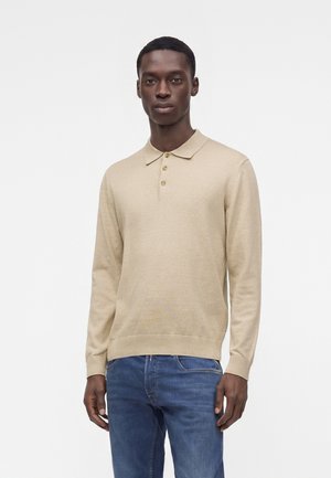 Homme portant un pull polo beige à manches longues et un jean bleu, debout devant un fond blanc uni.
