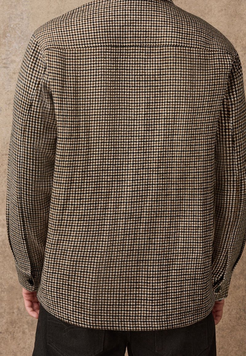 Vue de dos d'une personne portant une veste à motif pied-de-poule marron et noir avec des manches longues et un pantalon noir.