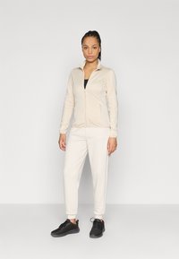 Φωτεινό μπεζ μπουφάν με φερμουάρ, εφαρμοστή σχεδίαση και γιακά, συνδυασμένο με ταιριαστά φωτεινά μπεζ τζόκερ και μαύρα αθλητικά παπούτσια. Μινιμαλιστικό στυλ.