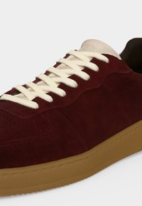 Gros plan d'une basket en daim bordeaux avec des lacets blancs, une languette beige et une semelle en caoutchouc sur un fond clair.