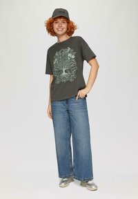 Donkergrijze t-shirt met een lichtgroene boomgraphic, gecombineerd met wijde blauwe jeans. Zilveren sneakers en een grijze pet maken de outfit compleet.