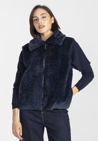 Kobieta w granatowej, futrzanej kamizelce zapinanej na zamek, nosząca długi rękaw i ciemne dżinsy, stoi z jedną ręką w kieszeni.