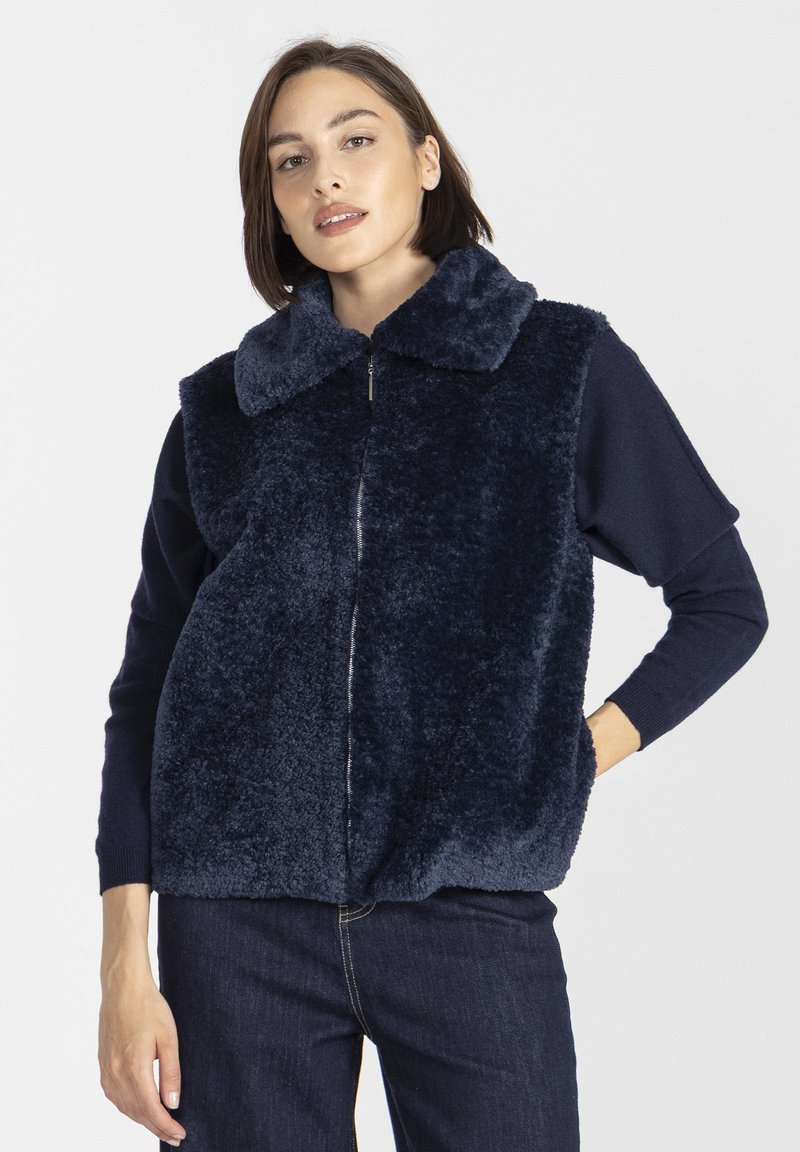 Kobieta w granatowej, futrzanej kamizelce zapinanej na zamek, nosząca długi rękaw i ciemne dżinsy, stoi z jedną ręką w kieszeni.