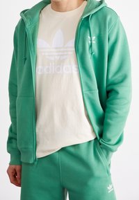 Felpa verde con zip realizzata in un tessuto morbido, con tasca frontale e lacci. Sotto, c'è una maglietta crema con un grande logo bianco.