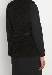 Czarna kamizelka z futra syntetycznego o fakturowanej, kudłatej powierzchni, przewiązana w talii gładkim czarnym paskiem, noszona na ciemnej bluzce z długim rękawem.