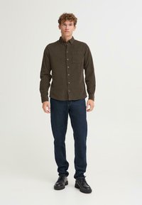 Camisa marrón de botones con un bolsillo, mangas largas y un tejido texturizado; combinada con jeans azul oscuro y calzado negro.