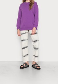 Sudadera de gran tamaño morada, pantalones blancos con patrón tie-dye negro, y sandalias negras con tachuelas. Estilo casual y cómodo.