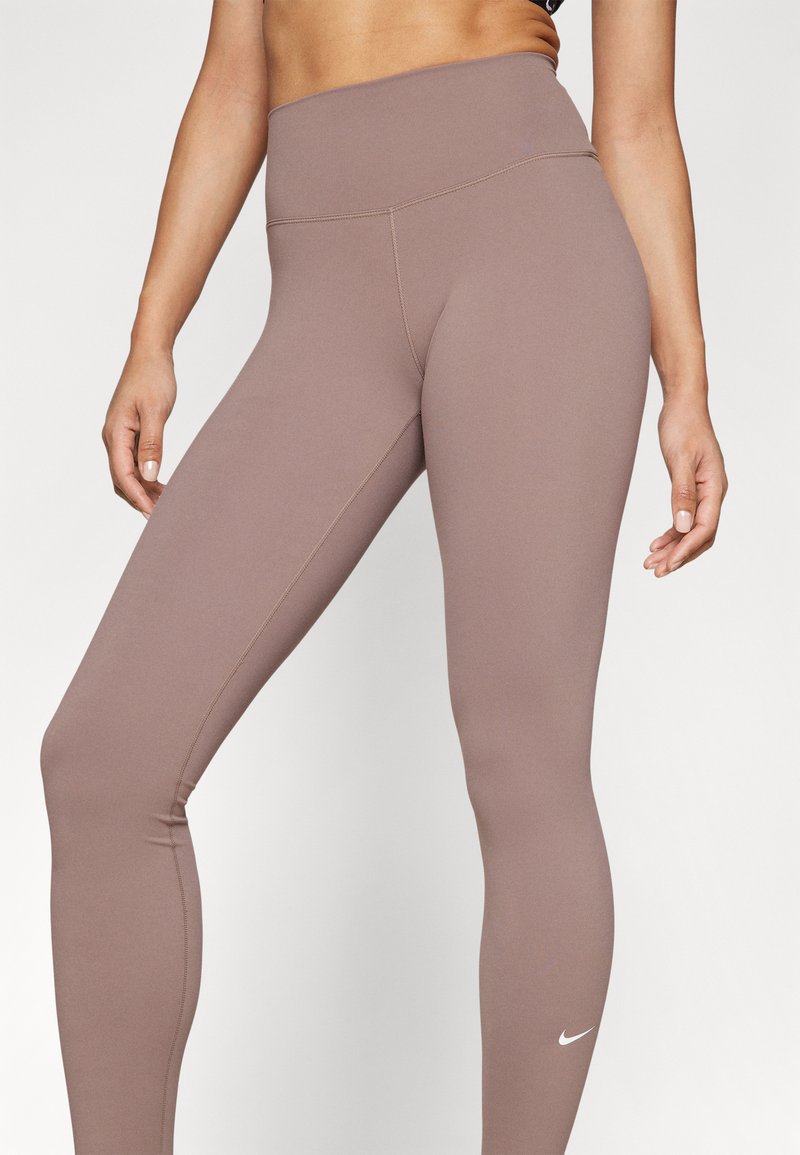 Hoge taille, figuur-hugging leggings in een gedempte bruine stof met een zachte textuur en een klein wit Nike-logo op het onderbeen.