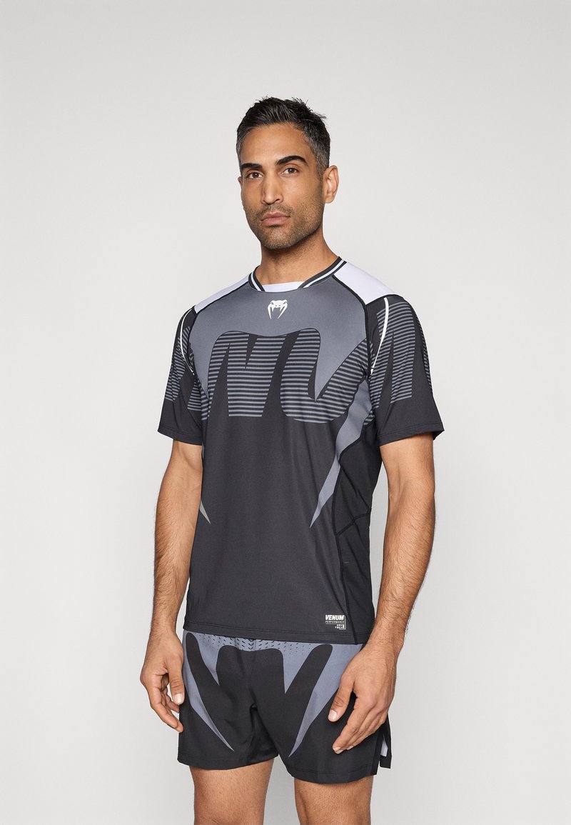 Camisola desportiva preta com padrões acentuados a cinzento e mangas curtas, combinada com calções a condizer, apresentando um logótipo e textura de tecido respirável.