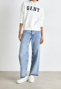Persona che indossa un hoodie bianco GANT, jeans azzurri a gamba larga con orli sfrangiati e sneakers platform bianche su uno sfondo neutro.