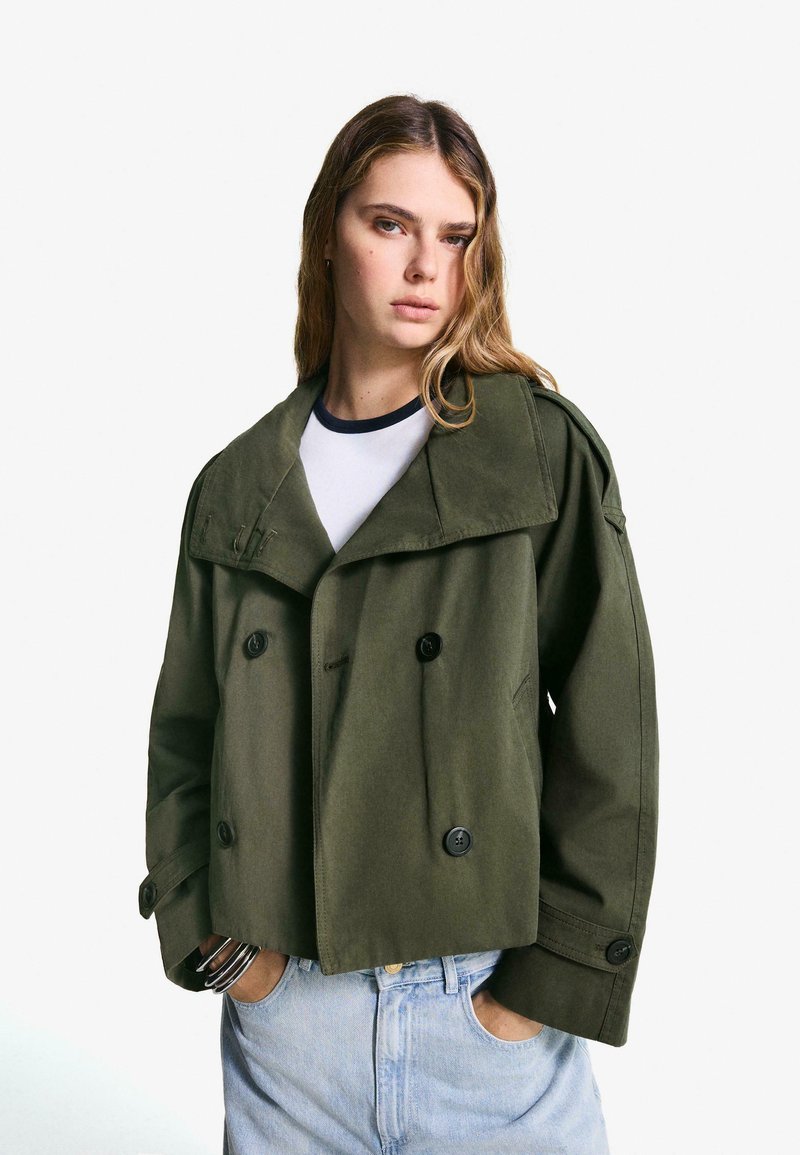 Kvinna med långt blont hår iförd en överdimensionerad olivgrön dubbelknäppt jacka, vit skjorta, ljusblå jeans och silvriga armband, med händerna i fickorna.