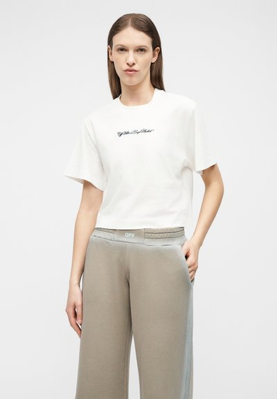 Γυναίκα με μακριά καστανά μαλλιά που φοράει λευκό cropped t-shirt με μαύρο κείμενο και μπεζ παντελόνι φαρδιάς γραμμής με λεπτομέρεια στη μέση που γράφει "OFF".