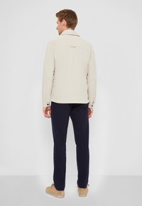 Giacca beige chiaro con una vestibilità morbida, caratterizzata da un colletto a punta e chiusure a bottone, abbinata a pantaloni blu navy e scarpe beige.