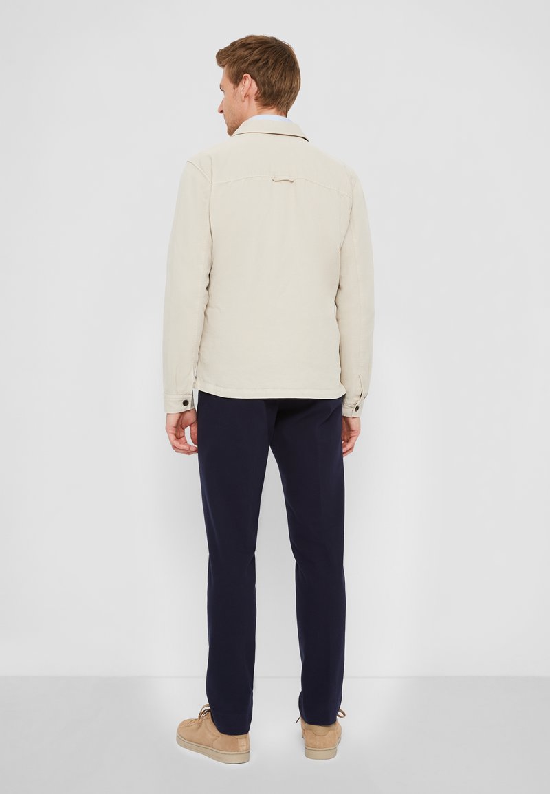 Hellbeige Jacke mit lockerer Passform, ausgestattet mit einem spitzen Kragen und Knopfverschluss, kombiniert mit navyfarbener Hose und braunen Schuhen.