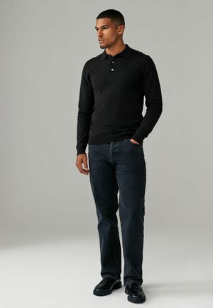 Sort langærmet polo sweater med krave og tre-knap lukning, parret med mørkeblå jeans og sorte sko, sat mod en ensfarvet baggrund.