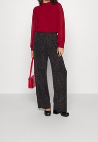 Blouse rouge à manches longues avec encolure froncée, accompagnée de pantalons noirs à large jambe texturés avec des détails scintillants argentés. Chaussures noires et sac à main rouge.