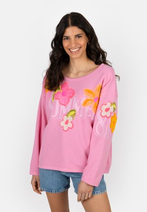 Young woman smiling, wearing pink long-sleeve shirt with colorful flower appliqués and blue denim shorts, standing against white background.