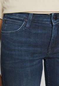 Tummansiniset farkut, joissa on näkyvä rakenne, viisi-taskumalli, kontrastiväriset ompeleet sekä metalliosat taskuissa ja napin sulkemisessa.