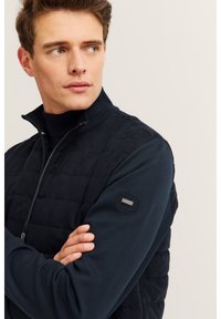 Man wearing a dark quilted zip-up jacket with hands crossed, looking to his left against a plain light background.