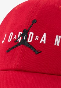 Culoare roșie, cu un design structurat, având broderie neagră a unei figuri sărind și cuvântul "JORDAN" în alb. Material din bumbac cu textură vizibilă.
