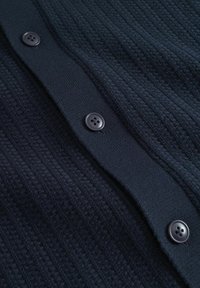 Marinblå stickad kofta med ribbat mönster, fem svarta knappar framtill och en slät krage.