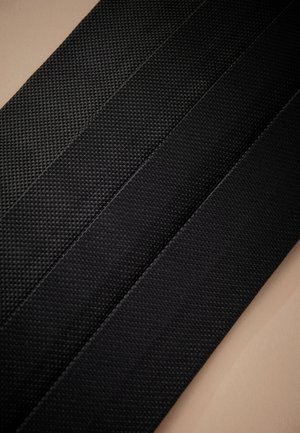 Vier schwarze, strukturierte Stoffriemen oder Gürtel, diagonal auf einem beigefarbenen Hintergrund angeordnet.