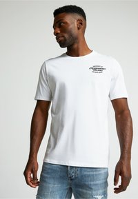 Valkoinen puuvillapaita, jossa on pyöreä kaula-aukko, lyhyet hihat ja musta graafinen teksti vasemmassa rinnassa. Yhdistetty vaaleansinisiin kuluneisiin farkkuihin.