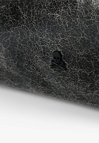 Superficie de cuero negro texturizado con acabado agrietado. Presenta un pequeño emblema de calavera y huesos cruzados en negro.